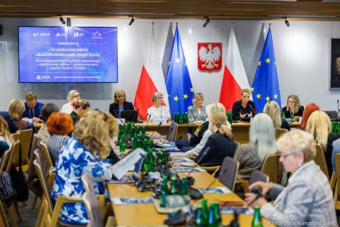 Kilkadziesiąt osób siedzi na sali przy stołach, pod jedną ze ścian flagi Polski i UE