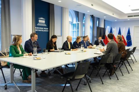 Dziesięć osób siedzi po dwóch stronach dużego stołu w sali konferencyjnej i rozmawia, w tle flagi Polski i UE i plansza z napisem "Rzecznik Praw Obywatelskich"