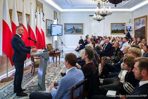 Kilkadziesiąt osób siedzi na sali i słucha wystąpienia osoby mówiącej za mównicą