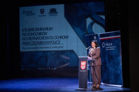 Osoba przemawia za mównicą na scenie w teatrze. W tle na dużym ekranie wyświetlony tytuł wydarzenia "VI Śląskie Seminarium Pełnomocników ds. Ochrony Praw Człowieka w Policji. Etyczne aspekty interwencji policyjnych"