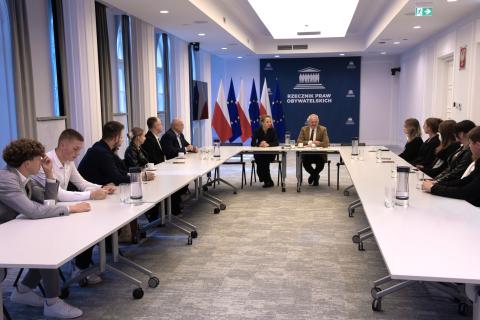 Kilkanaście osób siedzi przy stołach ustawionych w podkowę na sali konferencyjnej