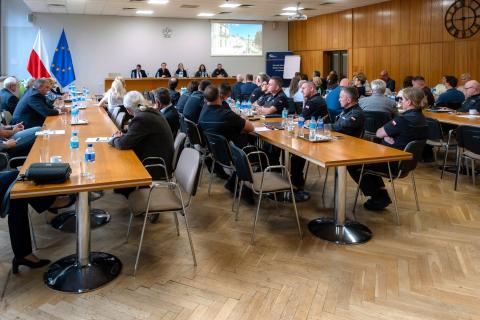 Kilkadziesiąt osób siedzi przy podłużnych stołach w dużej sali konferencyjnej