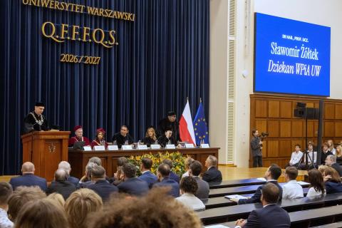Kilkadziesiąt osób siedzących na auli wykładowej słucha wystąpienia osoby mówiącej na scenie za mównicą ubranej w togę akademicką. Obok za stołem na scenie siedzi sześć osób również ubranych w togi akademickie.