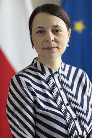 Portret Anny Błaszczak-Banasiak - lekko uśmiechnięta kobieta z krótkimi, czarnymi włosami na tle flag Polski i Unii Europejskiej