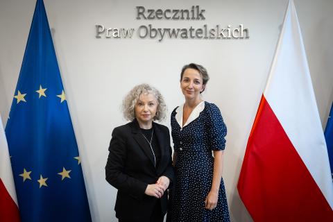 Sylwia Gregorczyk-Abram i Katarzyna Kotula uśmiechają się pozując do zdjęcia na tle flag Polski i Unii Europejskiej oraz napisu "Rzecznik Praw Obywatelskich"