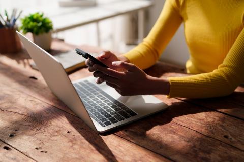 Kobieta trzymająca w dłoniach smartfona nad klawiaturą rozłożonego laptopa