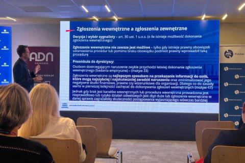 Osoba przemawia na sali konferencyjnej patrząc się na duży ekran z wyświetloną prezentacją
