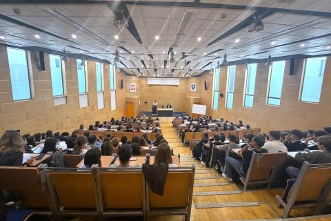 Kilkadziesiąt osób siedzi na dużej, uniwersyteckiej auli wykładowej i słucha wystąpienia osoby mówiącej na scenie za mównicą