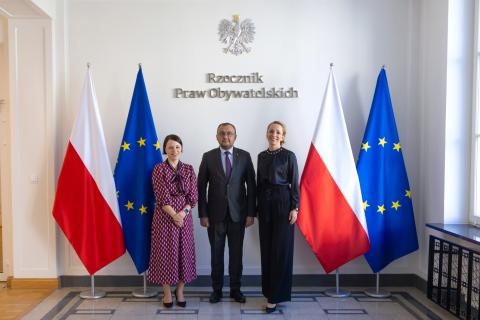 Trzy osoby pozują na stojąco do wspólnego zdjęcia na tle ściany z napisem "Rzecznik Praw Obywatelskich" i flag Polski i UE