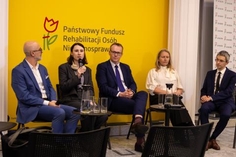 Pięć osób siedzi na krzesłach na sali konferencyjnej, jedna z nich mówi do trzymanego w dłoni mikrofonu