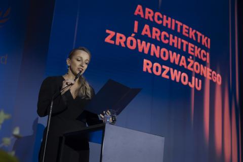 Osoba przemawia stojąc za mównicą na tle dużej planszy z napisem "Architektki i Architekci Zrównoważonego Rozwoju"