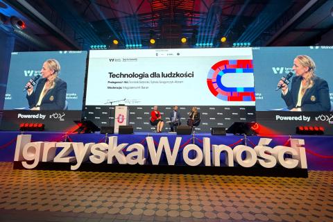Trzy osoby siedzą na fotelach na dużej scenie przed którą widoczny jest duży napis "Igrzyska Wolności", w tle na dużym wyświetlaczu zbliżenie na jedną z osób i grafika z napisem "Technologia dla ludzkości"