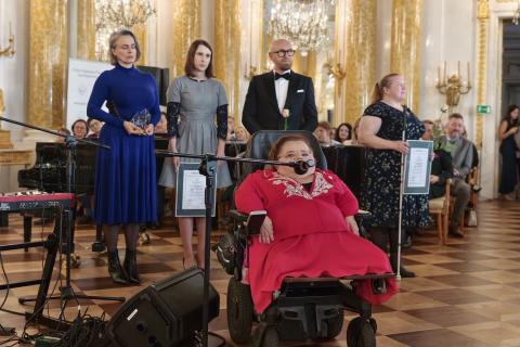 Osoba na wózku mówi do mikrofonu, za nią stoją cztery osoby i trzymają statuetki i oprawione dyplomy