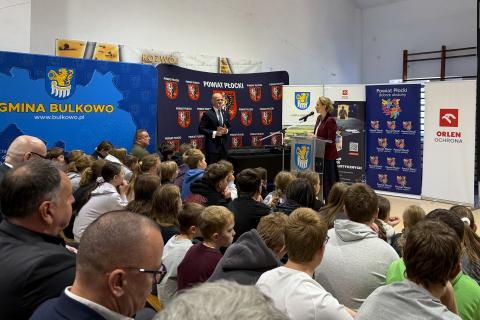 Osoba przemawia za mównicą na sali gimnastycznej w szkole, na widowni wystąpienia słuchają dzieci i dorośli