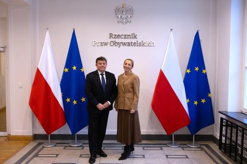 Dwie osoby pozują na stojąco do wspólnego zdjęcia na tle ściany z napisem "Rzecznik Praw Obywatelskich" i flag Polski i UE