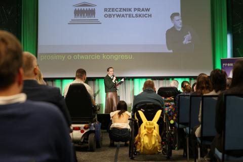 Osoba przemawia do ludzi zgromadzonych na sali konferencyjnej trzymając w dłoni mikrofon