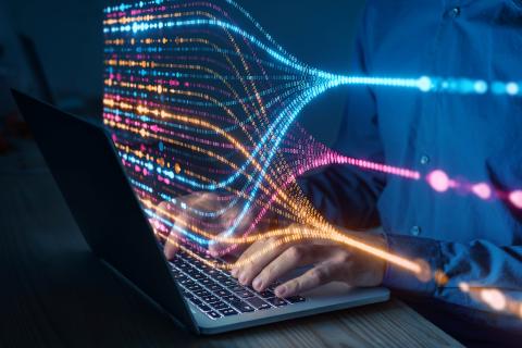 Osoba pisze na klawiaturze laptopa, ponad klawiaturą unosi się świecąca wizualizacja przepływu danych komputerowych