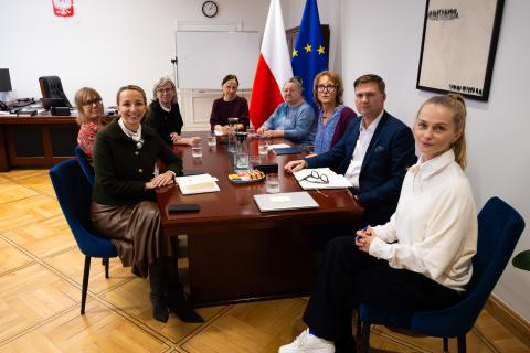 Osiem osób pozuje do zdjęcia siedząc wokół stołu konferencyjnego w gabinecie Rzecznika. W tle flagi Polski i Unii Europejskiej