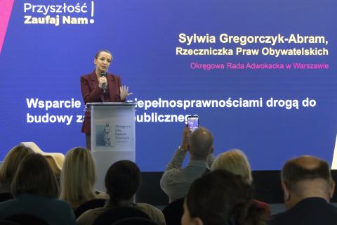 Sylwia Gregorczyk-Abram przemawia stojąc przy mównicy. Na pierwszym planie widać głowy osób z widowni