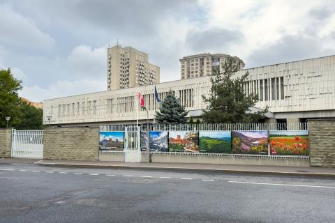 Kilkupiętrowy długi budynek widziany z boku otoczony ogrodzeniem, przed budynkiem na masztach flagi Polski i UE