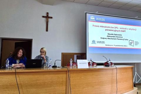 Dwie osoby siedzą za dużym stołem na sali konferencyjnej, jedna z nich trzyma mikrofon, za nimi w tle na wyświetlaczu widoczna prezentacja