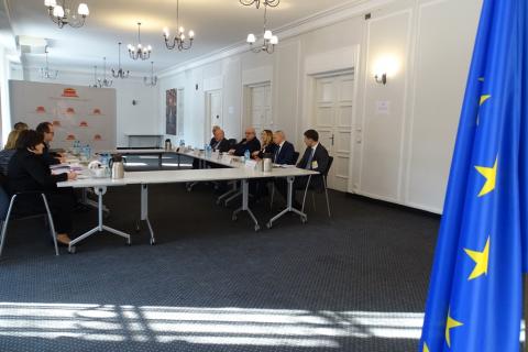 zdjęcie: po prawej stronie fragment flagi UE, w tle kilkanaście osób siedzi przy stole