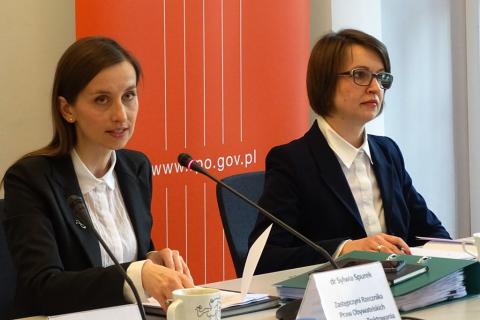 zdjęcie: dwie kobiety siedzą przy stole, koebieta po lewsj stronie mówi do mikrofonu, za nimi widać pomarańczowy baner