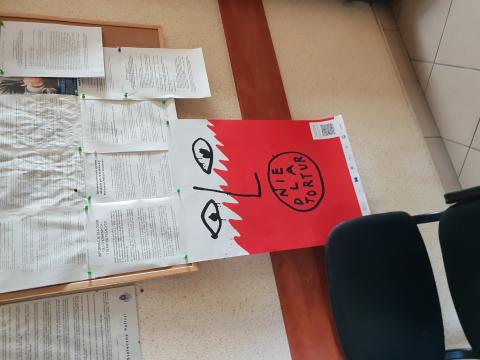 Na zdjęciu po lewej stronie widać krzesło, a nad nim znajduje się tablica korkowa, na której wisi plakat przedstawiający biało-czerwoną twarz z napisem: nie dla tortur