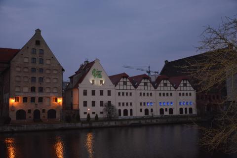 zdjęcie: z oddali widać stojący nad rzeką budynek, w którym okna zostały podświetlone na niebiesko