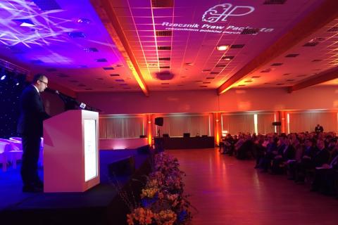 Mężczyzna przemawia w sali konferencyjnej oświetlonej czerwonymi i fioletowymi światłami