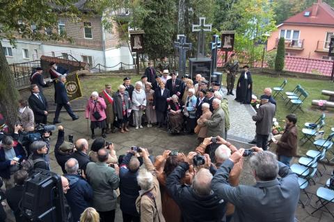 Zdjęcie: grupa starszych osób otoczona przez tłum. W tle domy i krzyże