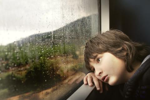 Zamyślony smutny chłopiec leżący na parapecie i wpatrujący się w deszcz za oknem.