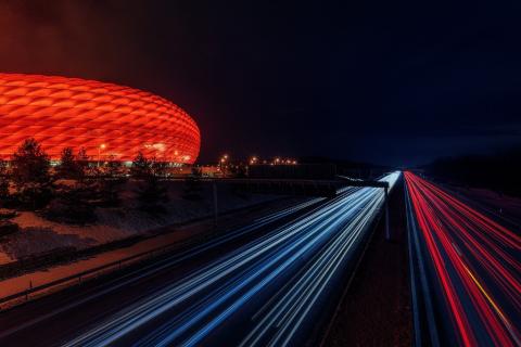 Samochody na szerokiej ulicy nocą, po lewej oświetlony na czerwono stadion piłkarski