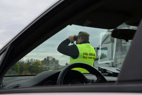 Funkcjonariusz Straży Granicznej w kamizelce odblaskowej stoi przed zaparkowanym na poboczu samochodem i obserwuje drogę przez lornetkę